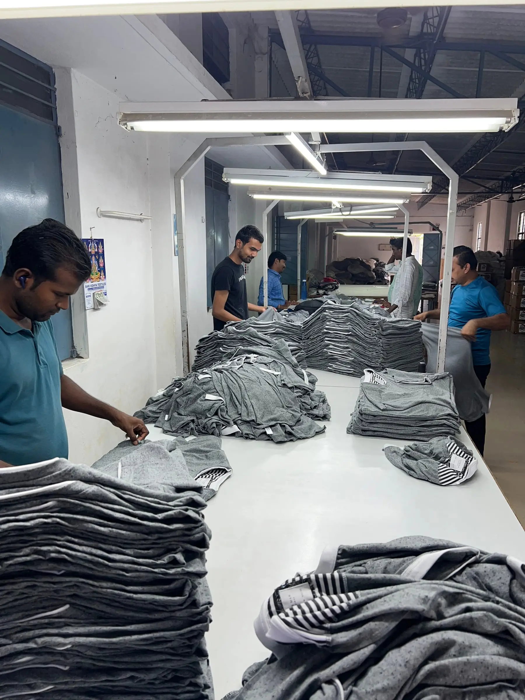 Folding and finishing line at the Tiruppur unit: stacks of grey t-shirts along a long white table, with the team folding and checking on both sides.
