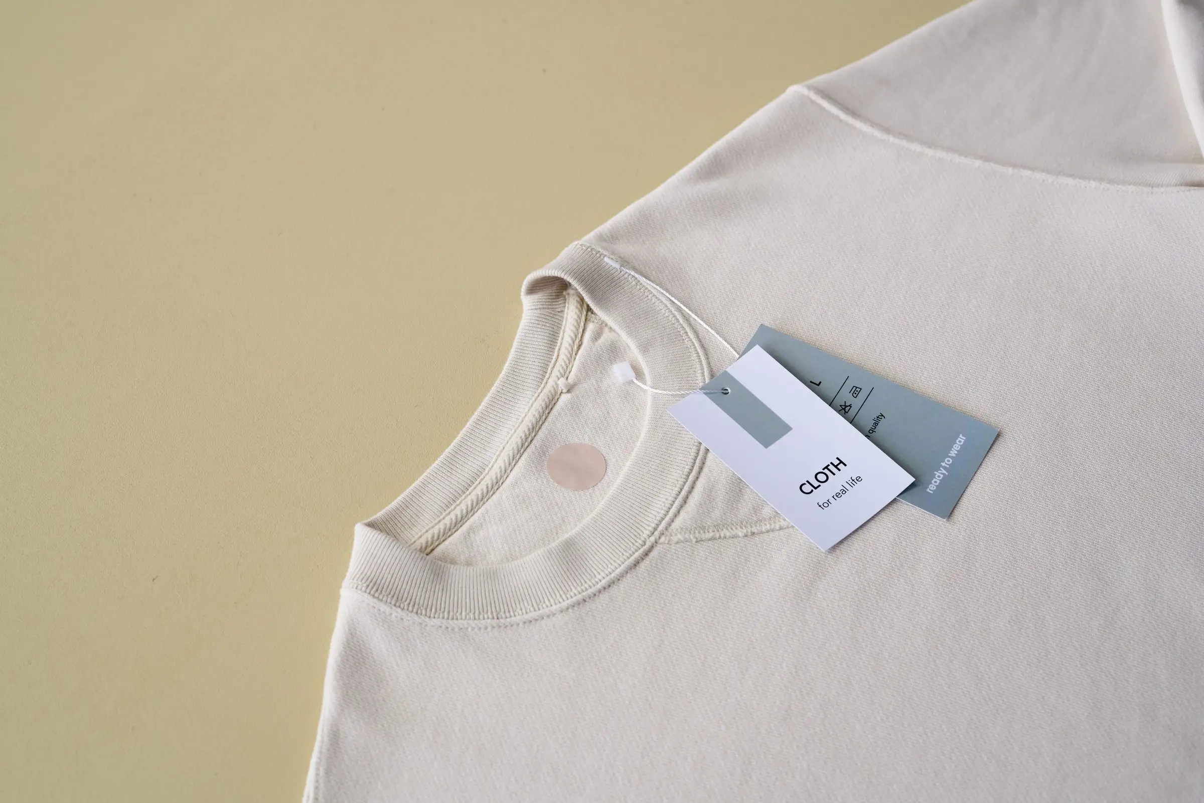 A finished natural-white crew-neck t-shirt laid flat on a pale yellow surface, a blank hang tag tied at the neckline.